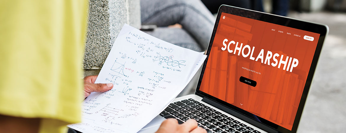 Someone using a laptop which has the word Scholarship displayed on it. The person is holding a piece of paper with formulas on it.